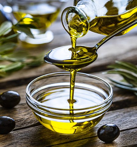 Pouring extra virgin olive oil from a spoon to a glass container. Olive branches comes from the left and right. Some olive oil bottles are out of focus at background. Black olives are on the table and complete the composition. The composition is on a rustic wooden kitchen table. Predominant colors are gold, green and brown. High resolution 42Mp studio digital capture taken with Sony A7rII and Sony FE 90mm f2.8 macro G OSS lens